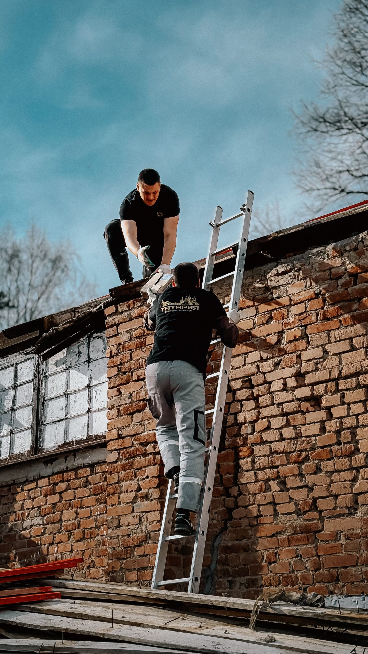 Two construction workers using a ladder to reach a brick building roof under a blue sky.